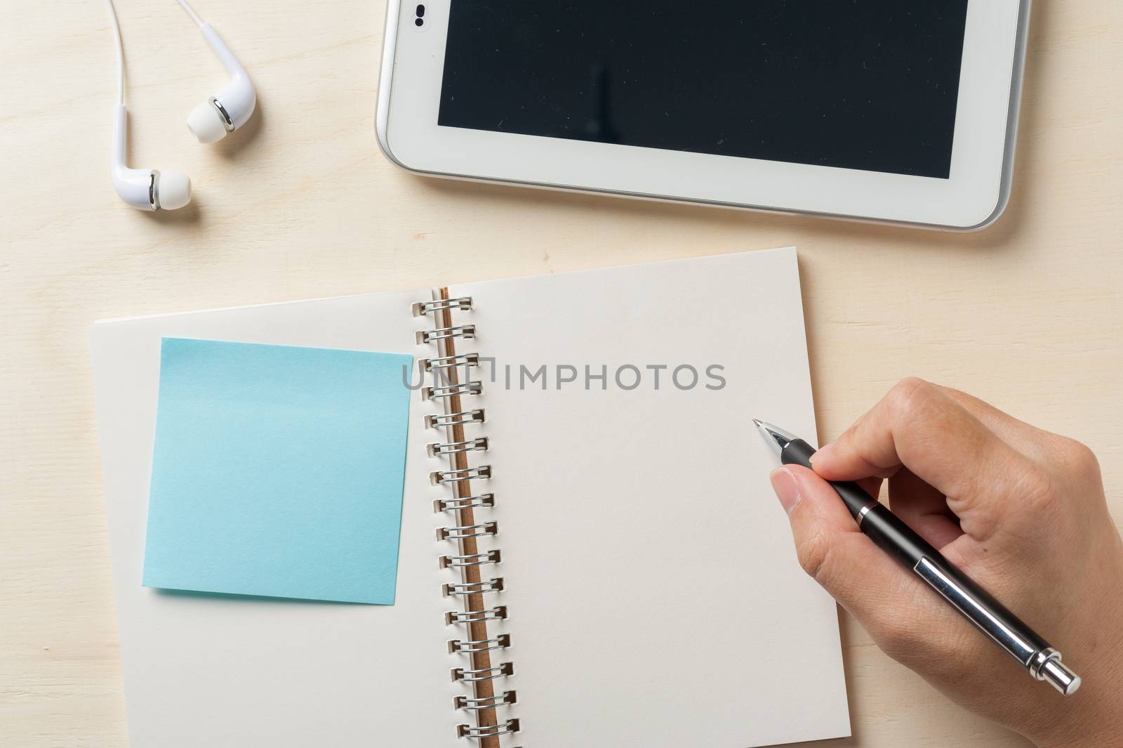 Young man right hand writing on notebook with sticky paper beside small tablet pc and in ear headset on wood table