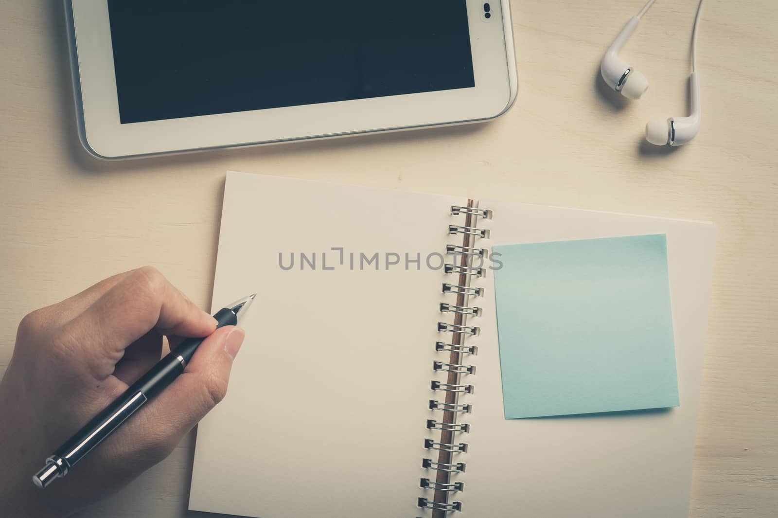 Young man left hand writing on notebook with sticky paper beside small tablet pc and in ear headset on wood table with vintage filter effect