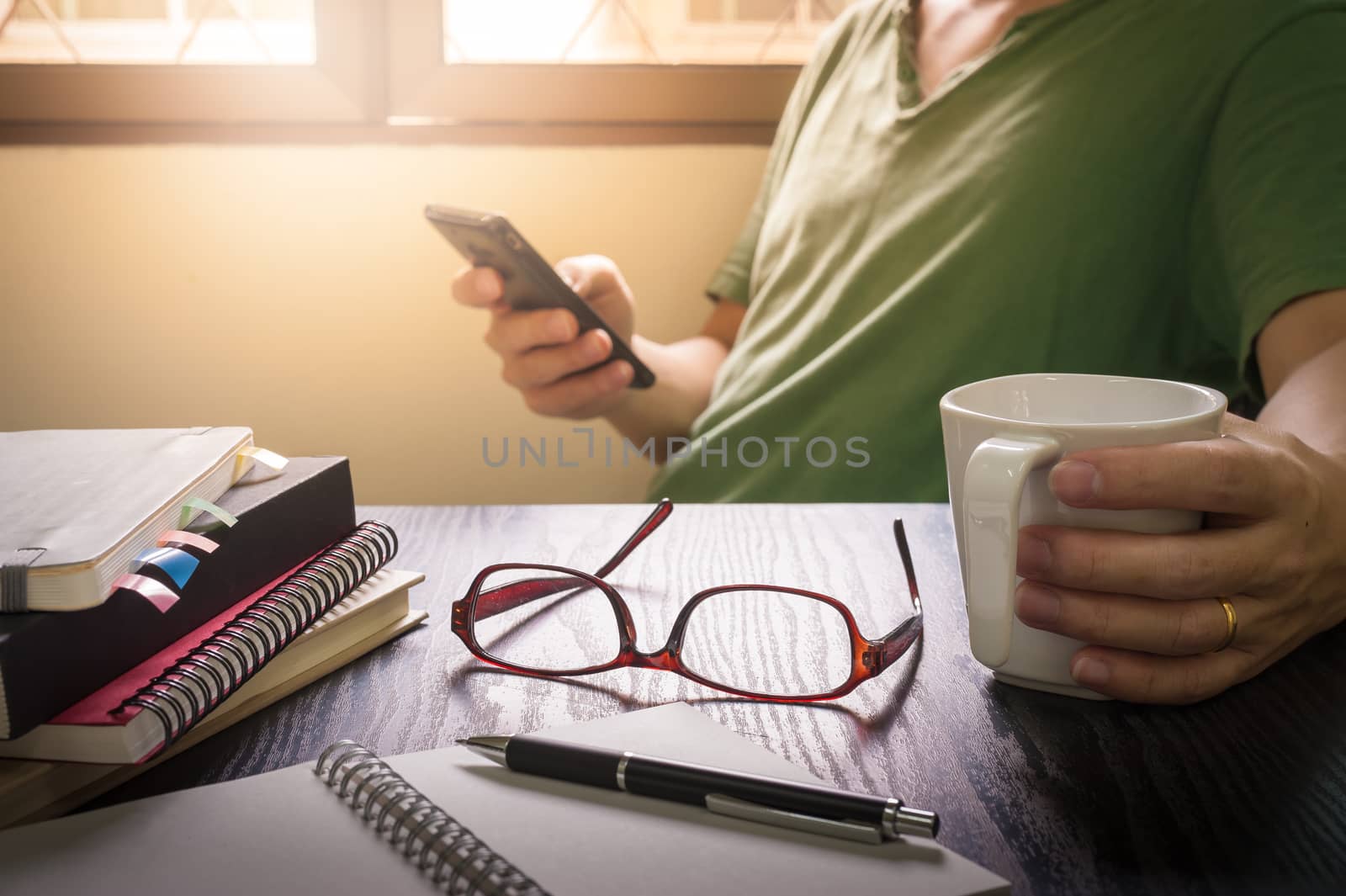 Glasses put down on table beside notebooks and pen while young freelance worker using smart phone and holding coffee cup in morning time on work day, Working at home concept