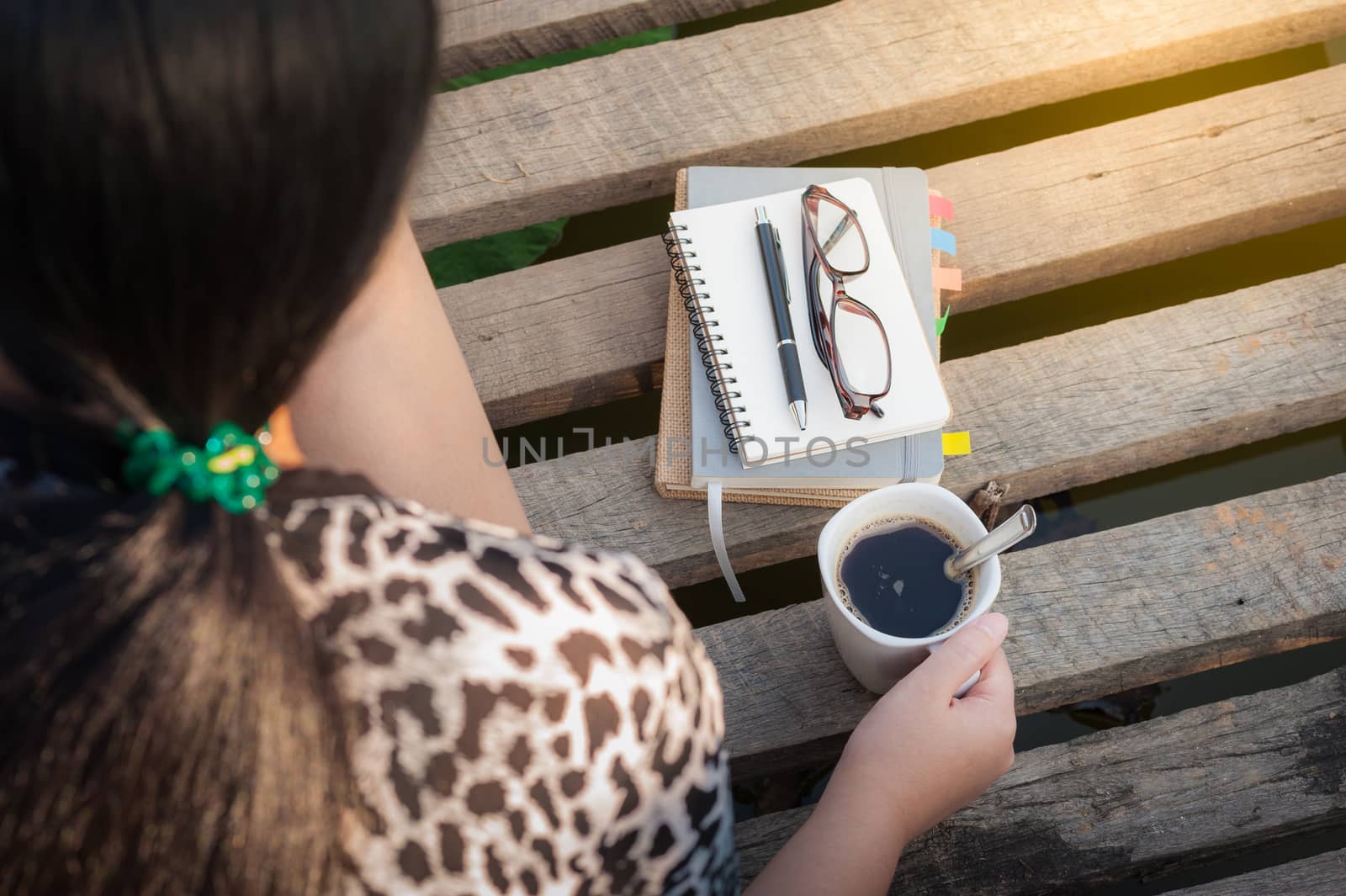 Young woman holding coffee cup while sitting on wood bridge with notebooks, pen, and glasses in morning time on weekend
