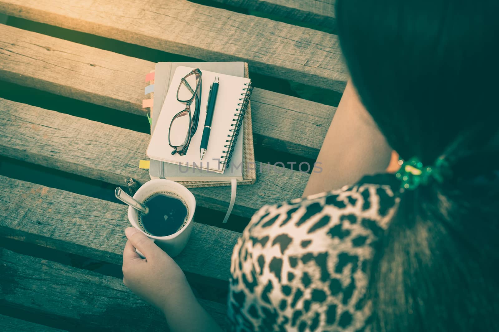 Young hipster woman holding coffee cup while sitting on wood bridge with notebooks, pen, and glasses in morning time on weekend with vintage filter effect