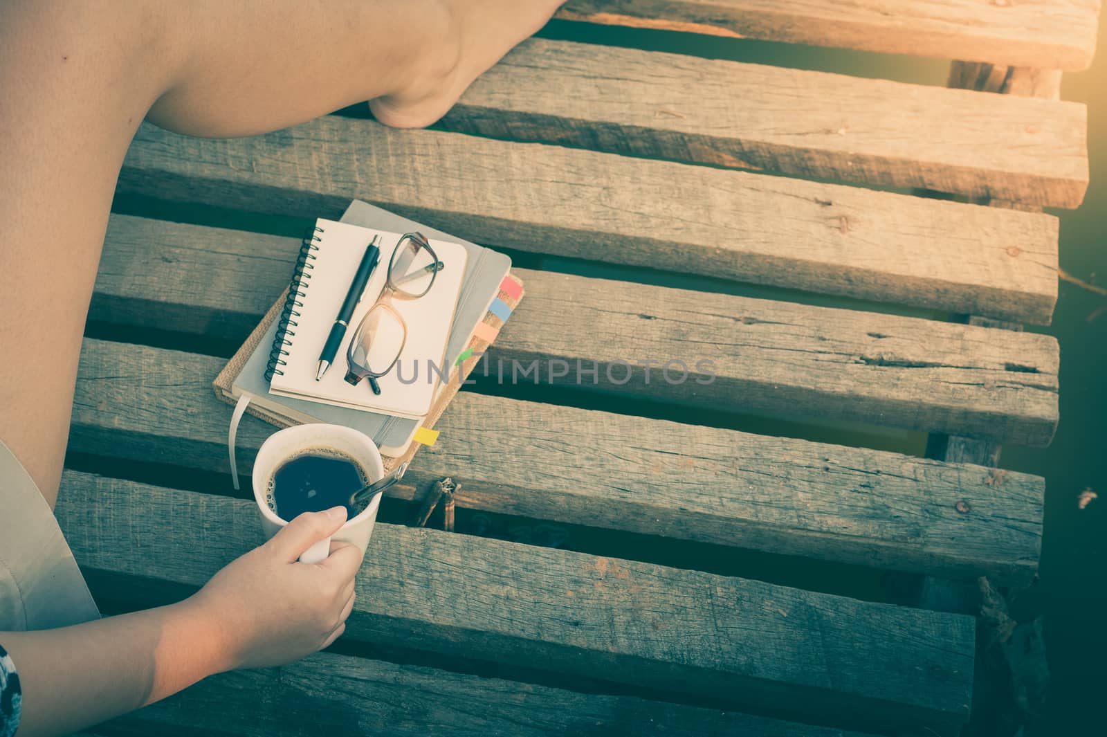 Young hipster woman holding coffee cup on wood bridge with notebooks, pen, glasses in morning time on weekend with vintage filter effect