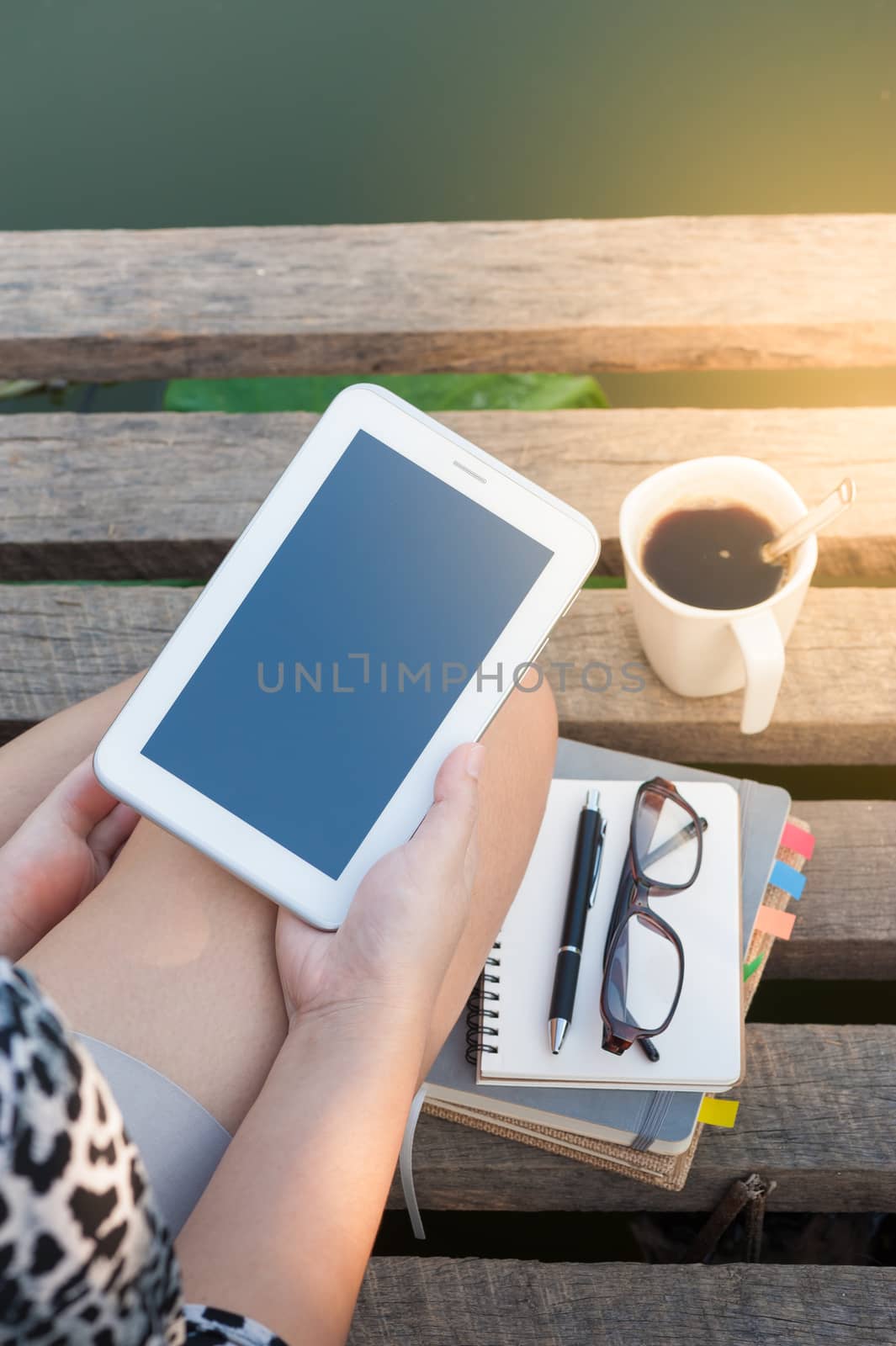 Young woman holding small talet with right hand while sitting on wood bridge with coffee cup, glasses, and notebooks in morning time on weekend
