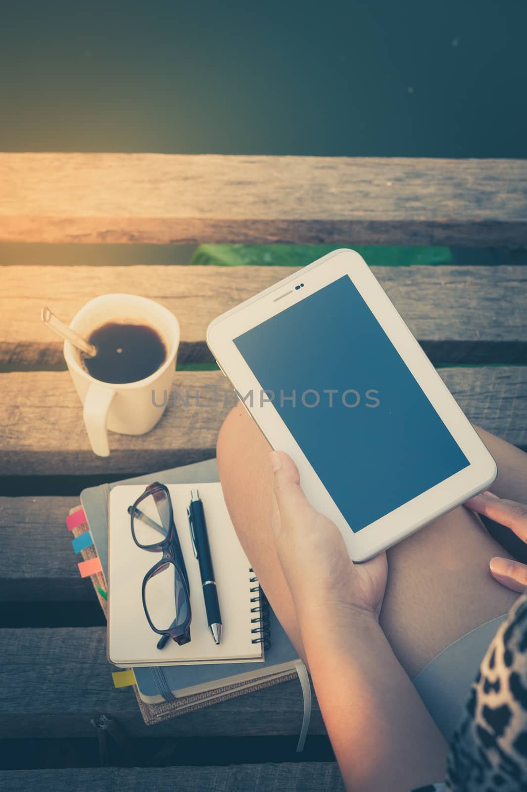 Young woman holding small talet with left hand while sitting on wood bridge with coffee cup, glasses, and notebooks in morning time on weekend with vintage filter effect