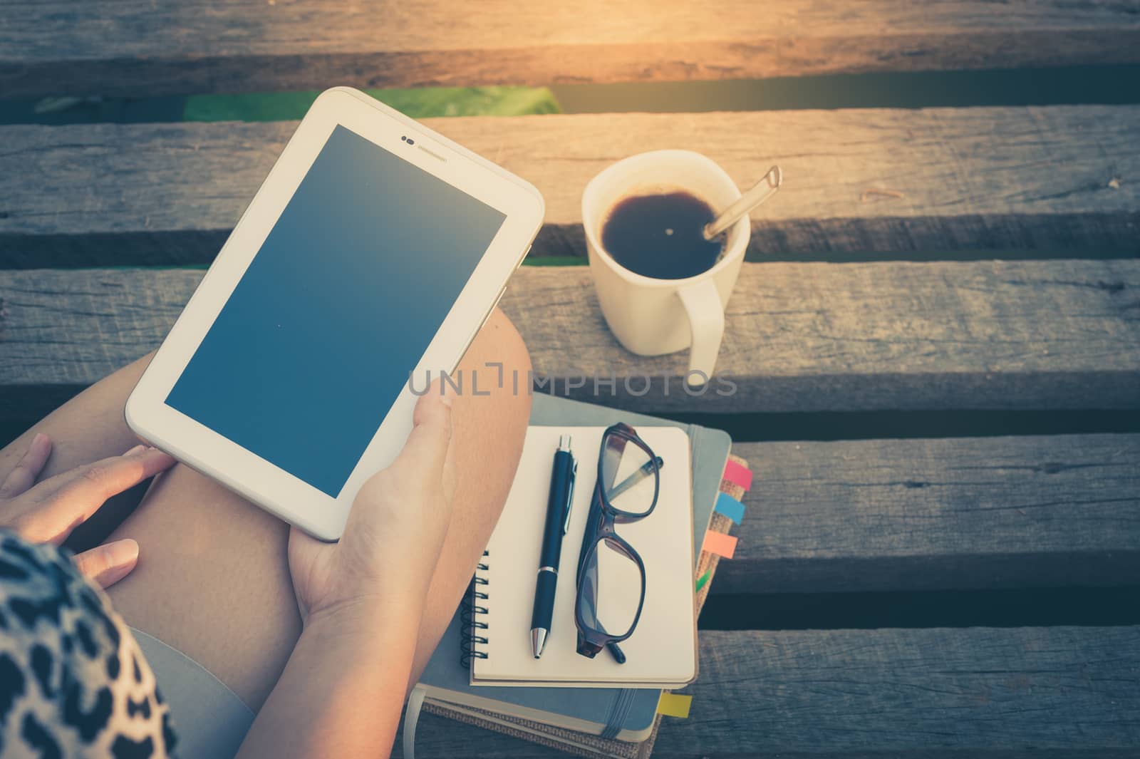 Young woman holding small talet while sitting on wood bridge with coffee cup, glasses, and notebooks in morning time on weekend with vintage filter effect