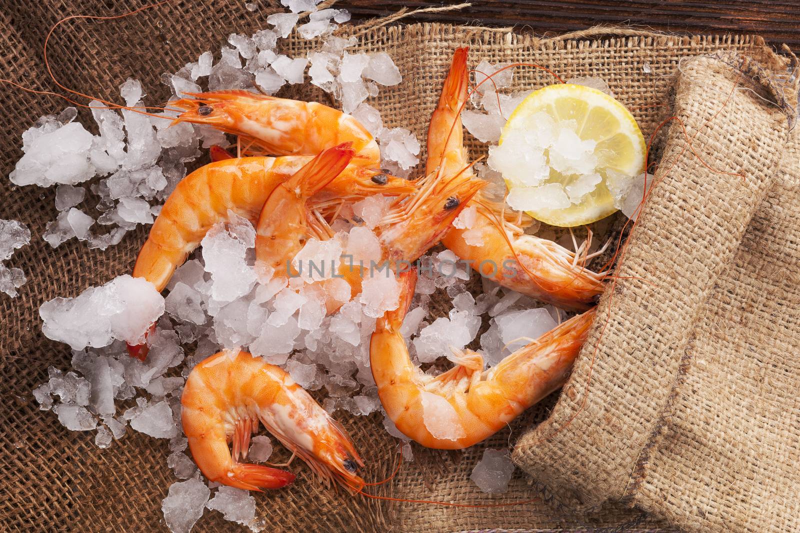 Fresh shrimp with lemon on ice in brown burlap bag on wooden table, top view. Culinary seafood eating.