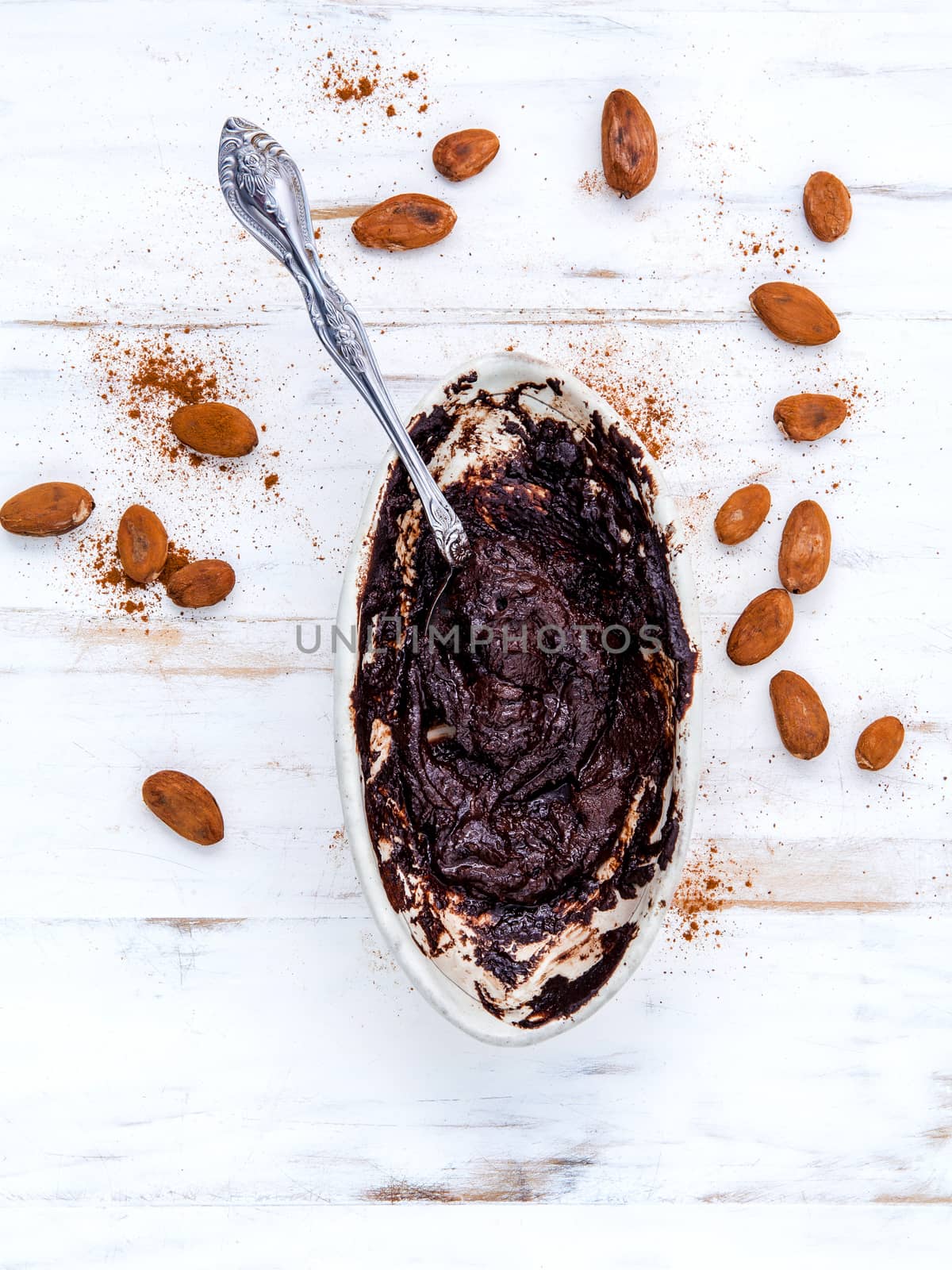 Melted chocolate with metal spoon in ceramic bowl and roast cocoa beans  on white table.