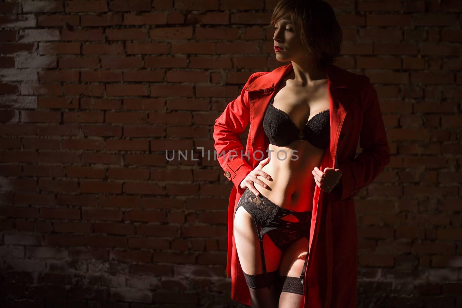 Attractive young woman alluring in sexual lingerie and red coat at grunge industrial setting. Beauty, fashion. Concept: seduction, exhibitionism.