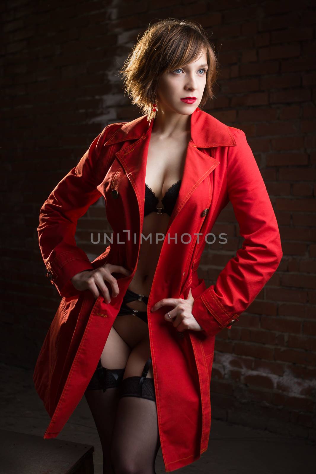 Attractive young woman alluring in sexual lingerie and red coat at grunge industrial setting. Beauty, fashion. Concept: seduction, exhibitionism.