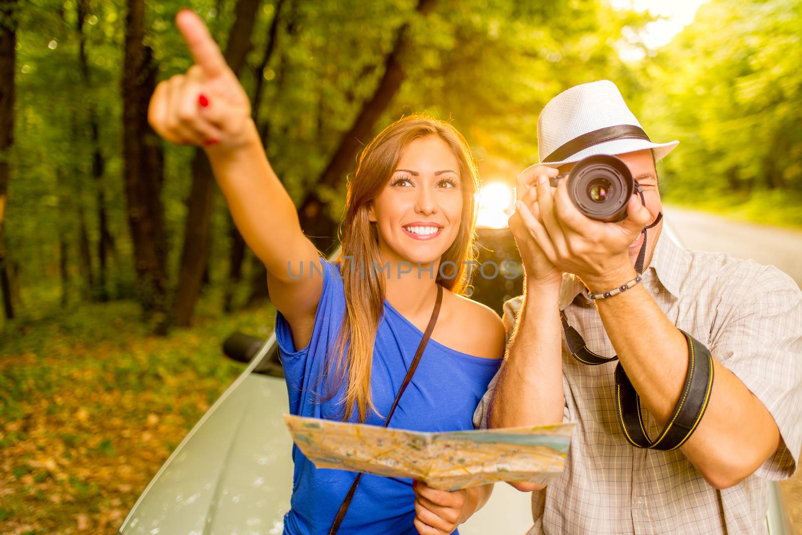 Young travelers standing before a car in the forest. Girl holding map and pointing destination. Guy taking a photo with digital camera.