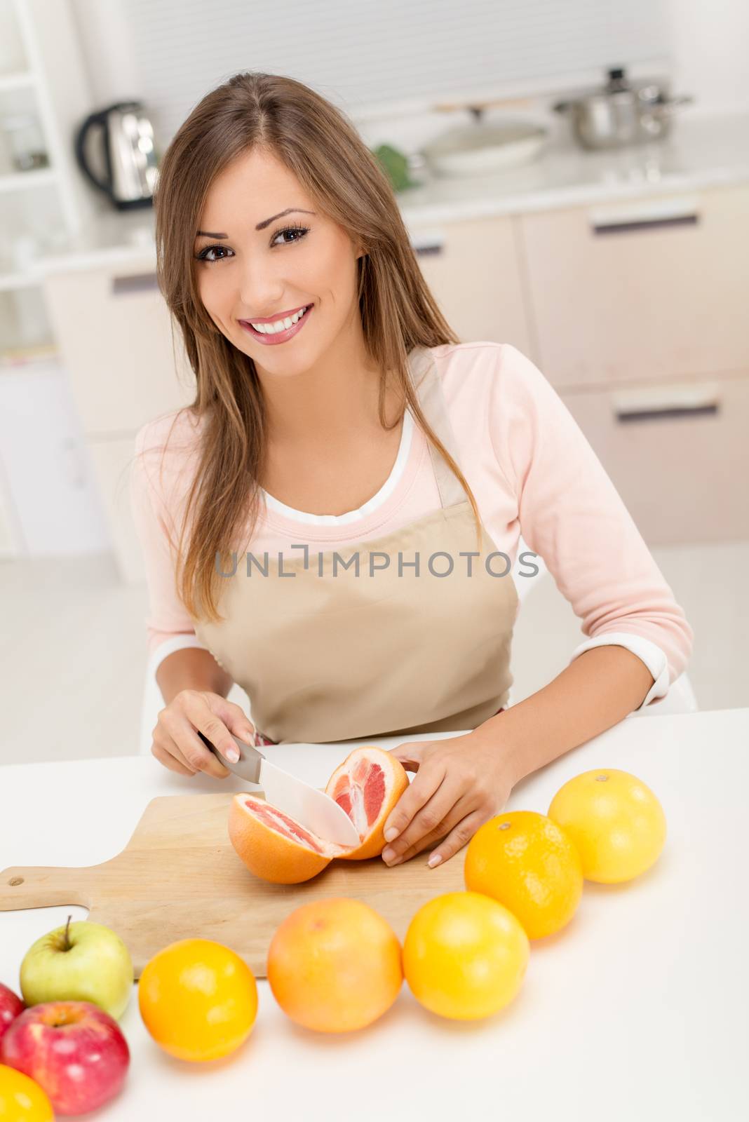 Cute young woman cutting grapefruit in kitchen. Looking at camera.