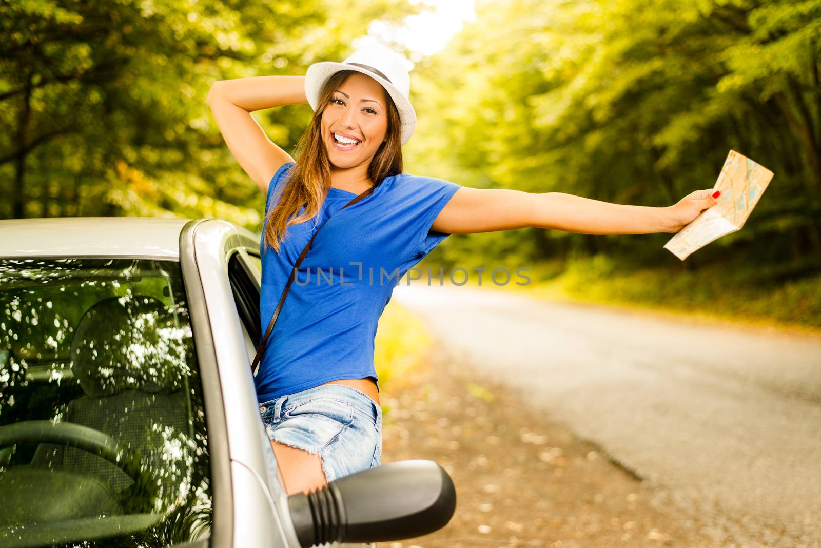 Cheerful woman on summer travel vacation leaning out of a car window. She is holding map with arms raised.