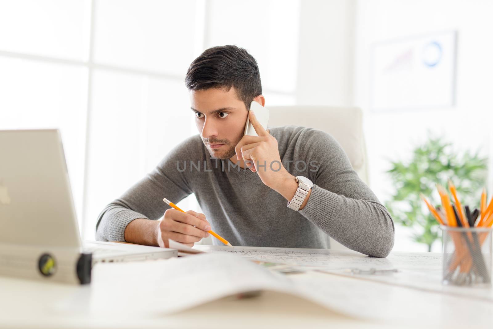 Young arabian male contractor sitting at desk in office, using phone and analyzing blueprint. He looking at the laptop in front him.