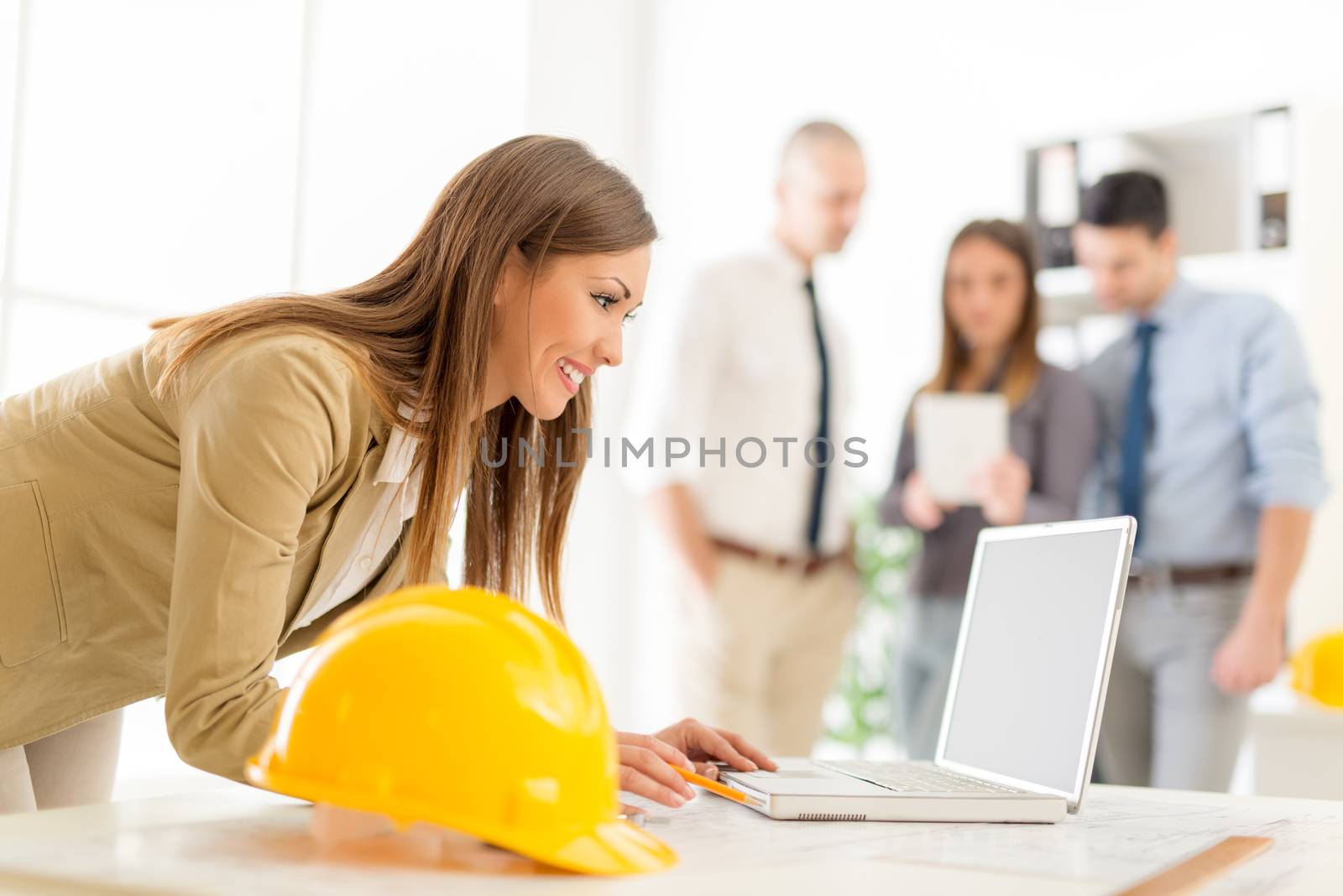 Smiling young female architect analyzing project at laptop in office. Her colleagues standing in background. 