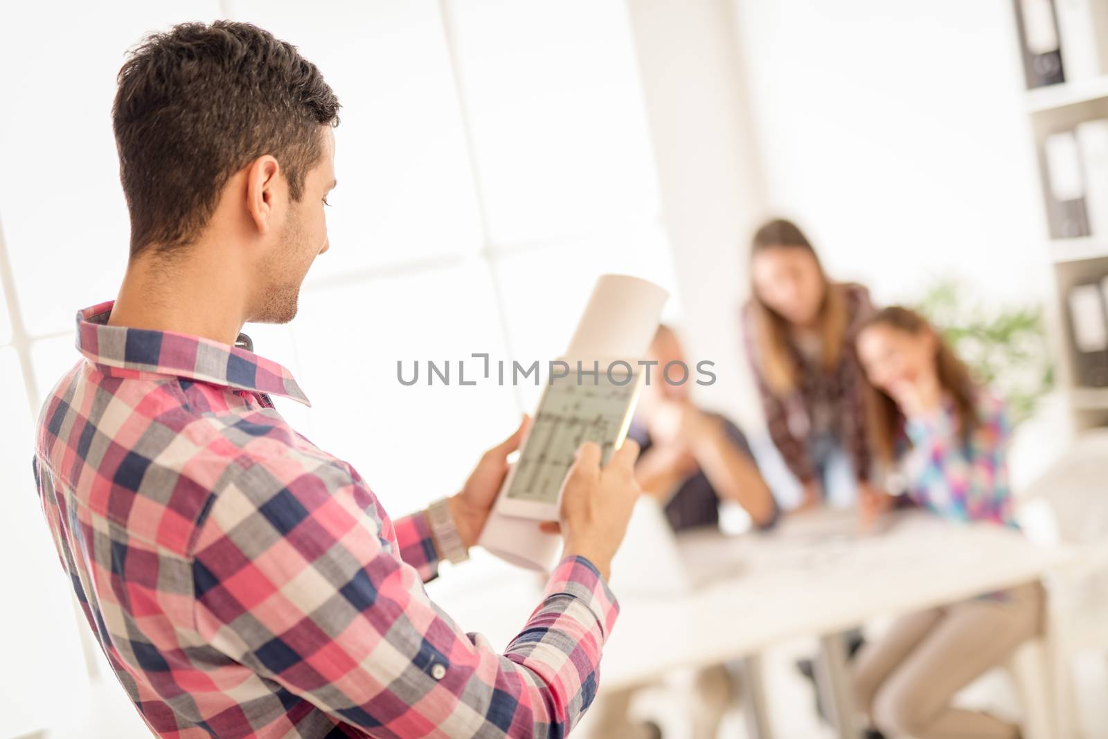 Young arabian male designer checking the architectural plans on a tablet
in office. His colleagues sitting in background. 