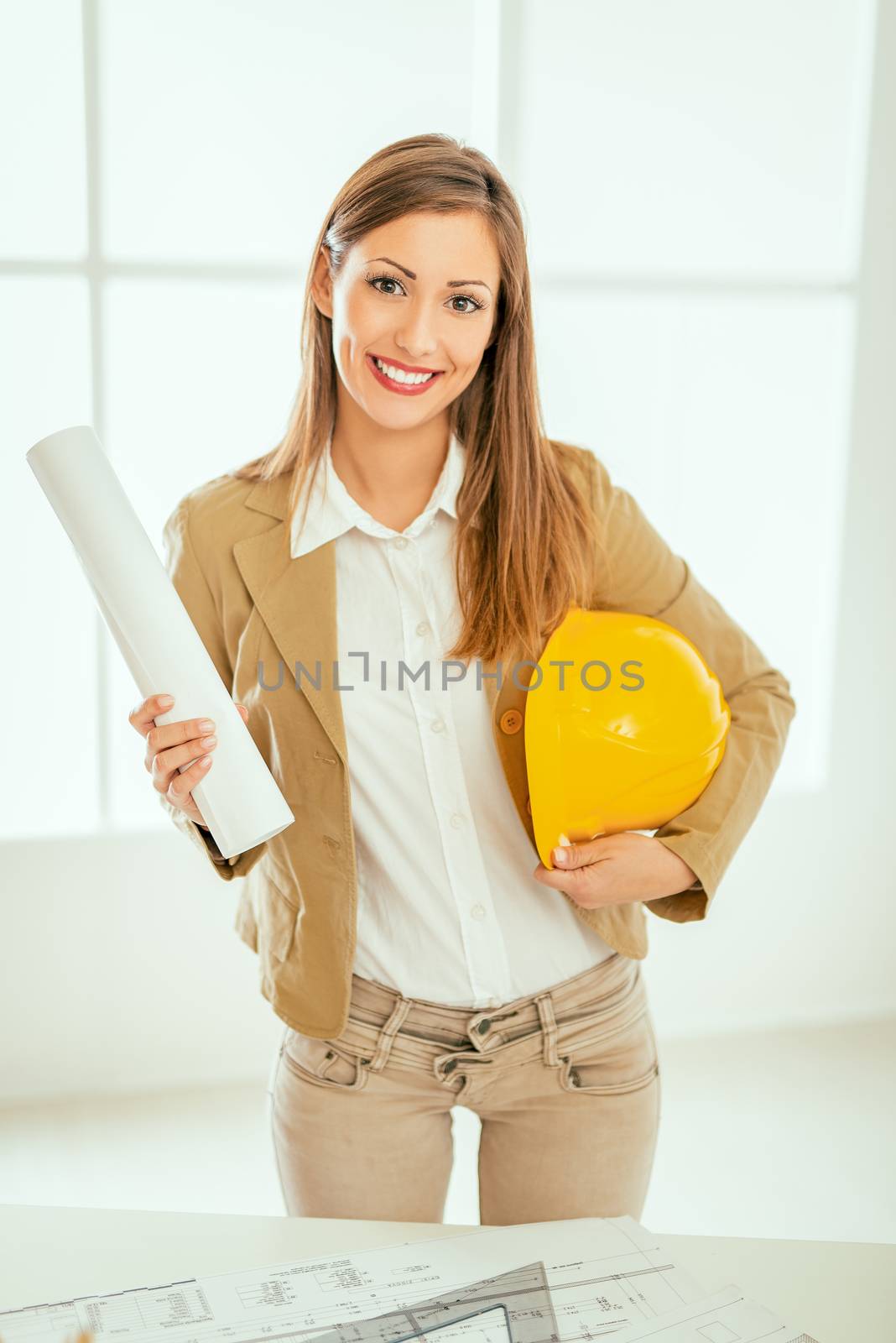 Young successful female architect standing in the office and holding helmet and project. Looking at camera.