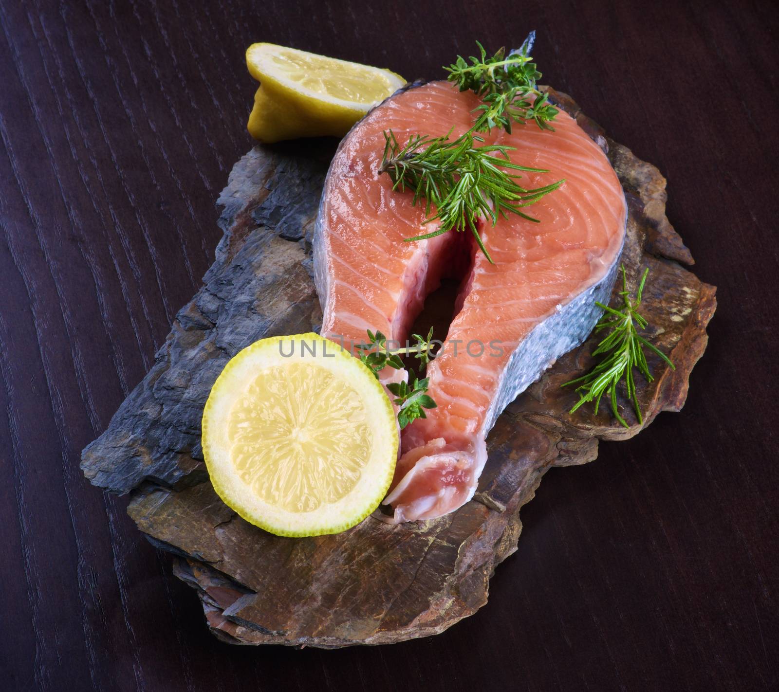 Perfect Raw Salmon Steak with Slices of Lemon and Rosemary on Shale Stone Board closeup on Dark Wooden background