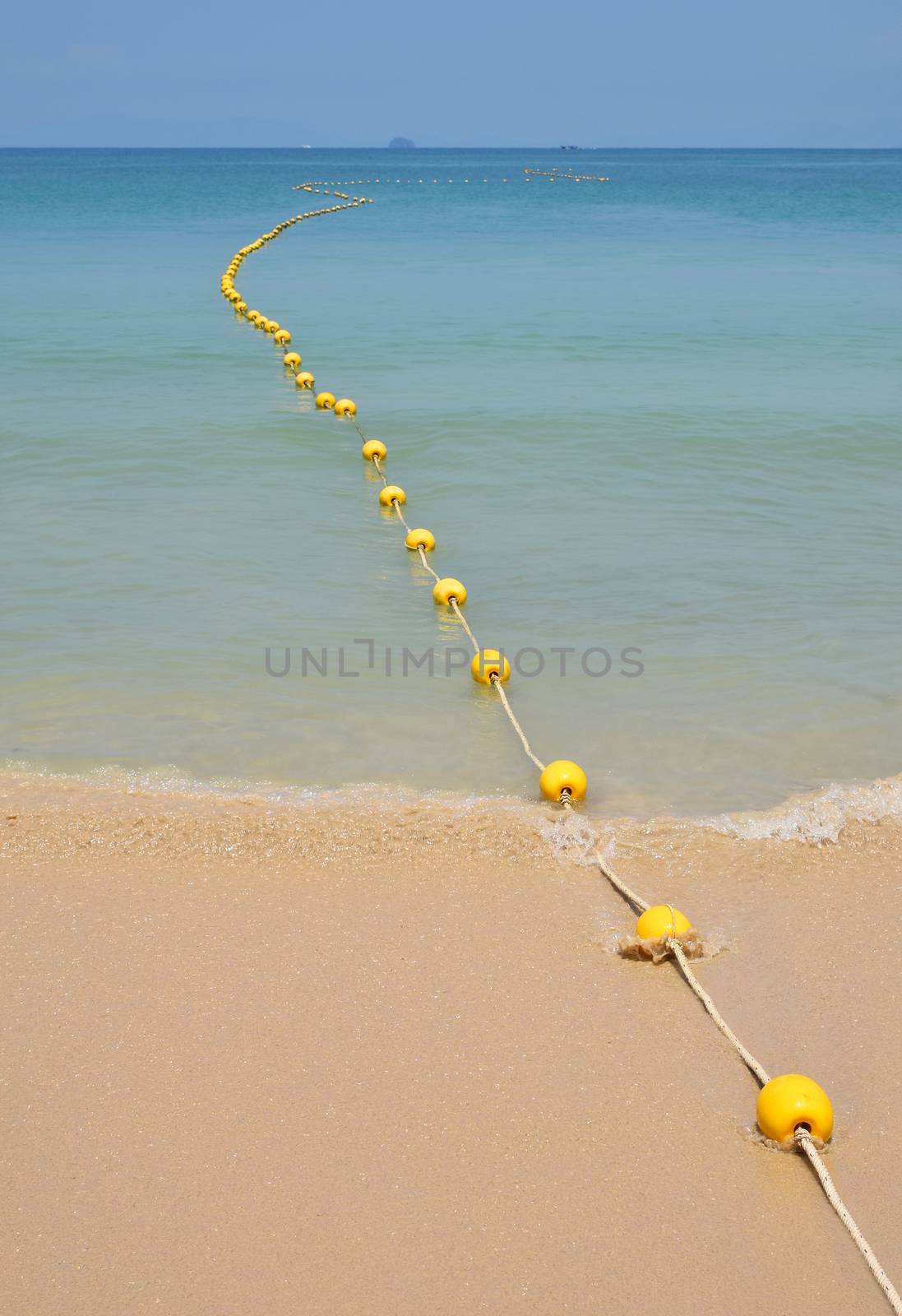 Chain of yellow polystyrene sea marker buoys with cable tow at sand beach and in blue sea water with clear sky above, perspective