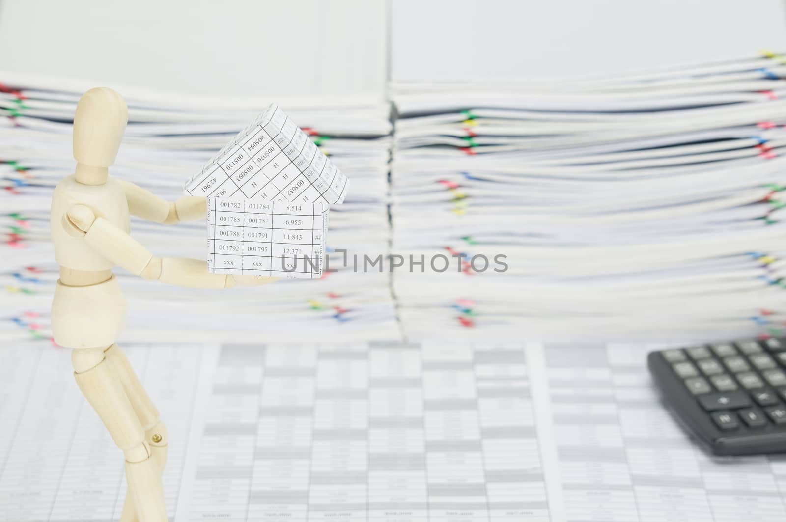 Wooden dummy holding house on finance account have blur calculator and overload of paperwork as background.