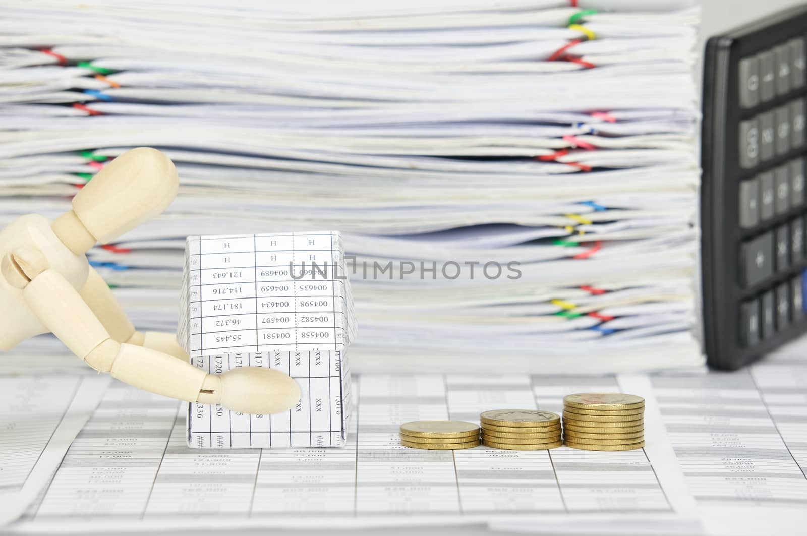 Wooden dummy holding house and step pile of gold coins on finance account have blur calculator place vertical with overload of paperwork as background.