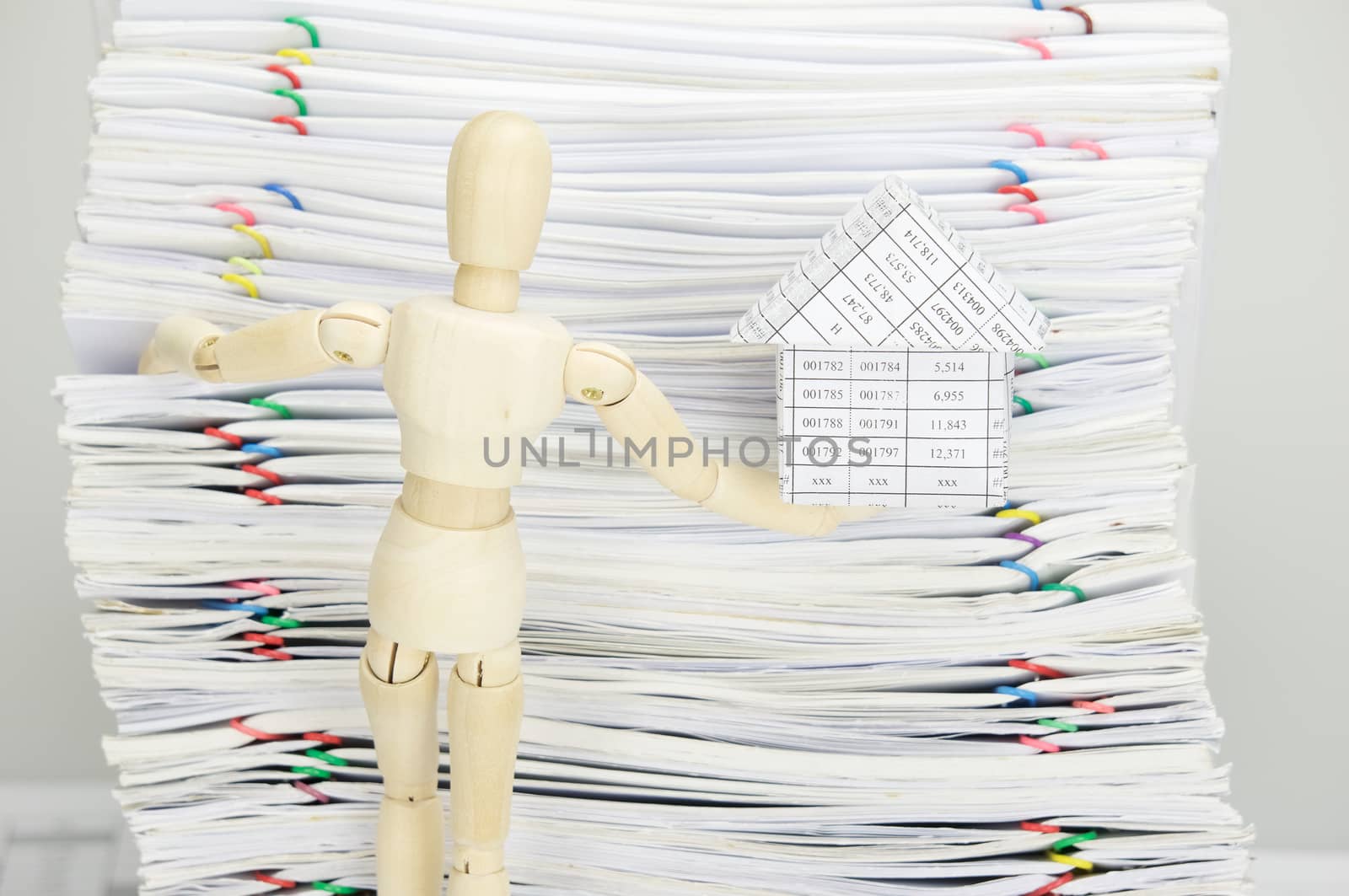 Wooden dummy holding house and climb on overload of paperwork with colorful paperclip as background.