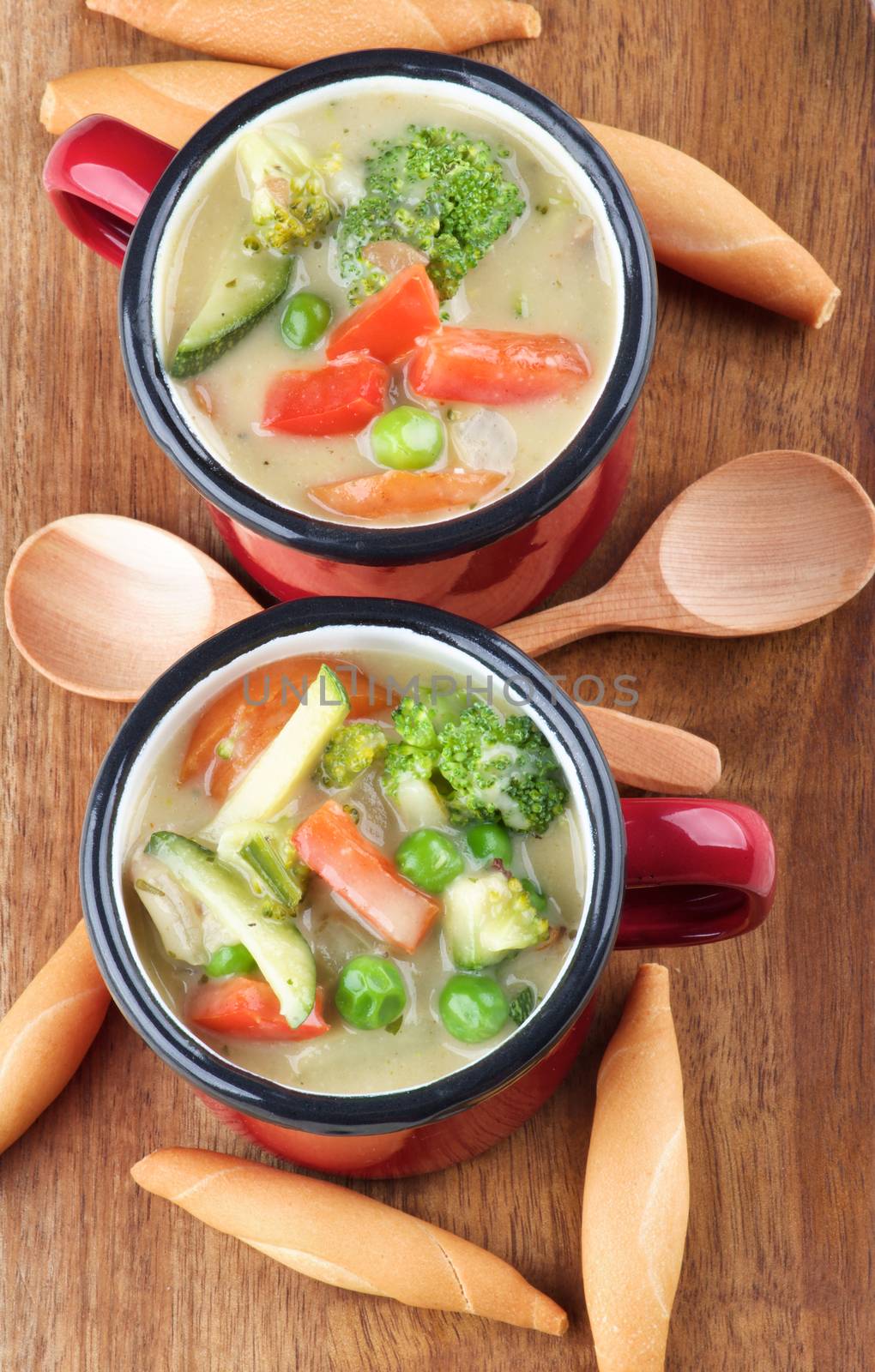Delicious Homemade Vegetables Creamy Soup with Broccoli, Carrots, Zucchini, Leek, Red Bell Pepper and Green Pea in Red Soup Cups with Wooden Spoon and Bread Sticks closeup on Wooden Cutting Board