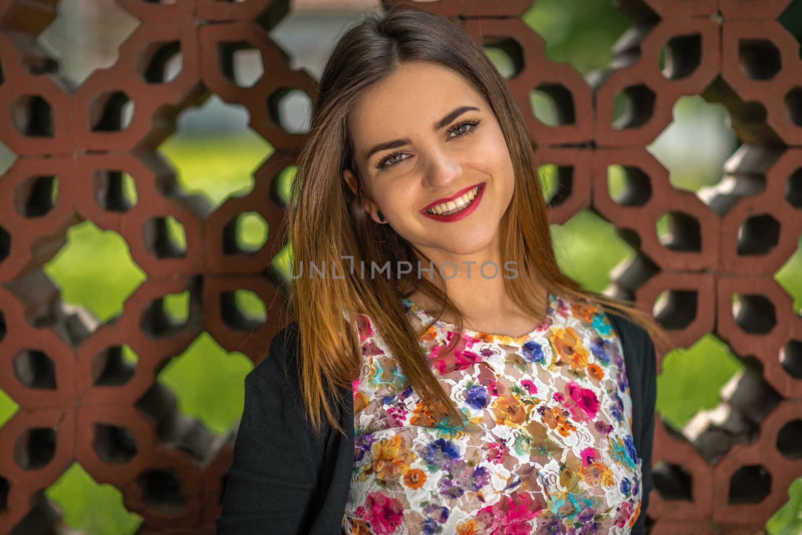 Portrait of a beautiful young woman with big smile, bright eyes and red lips against a fence made from ornamental bricks. Shallow depth of field.