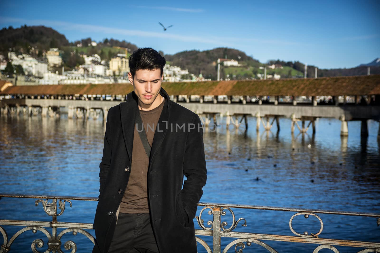 Handsome man standing near metal fence in Lausanne. Famous wooden bridge and Water Tower on background.