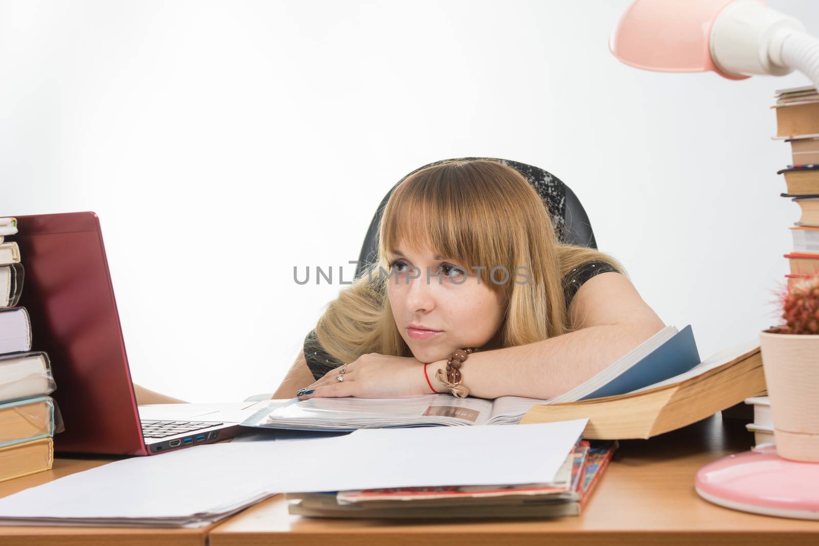 The girl at the desk crammed with books and papers sadly looks at the laptop with her head resting on his hand on the table