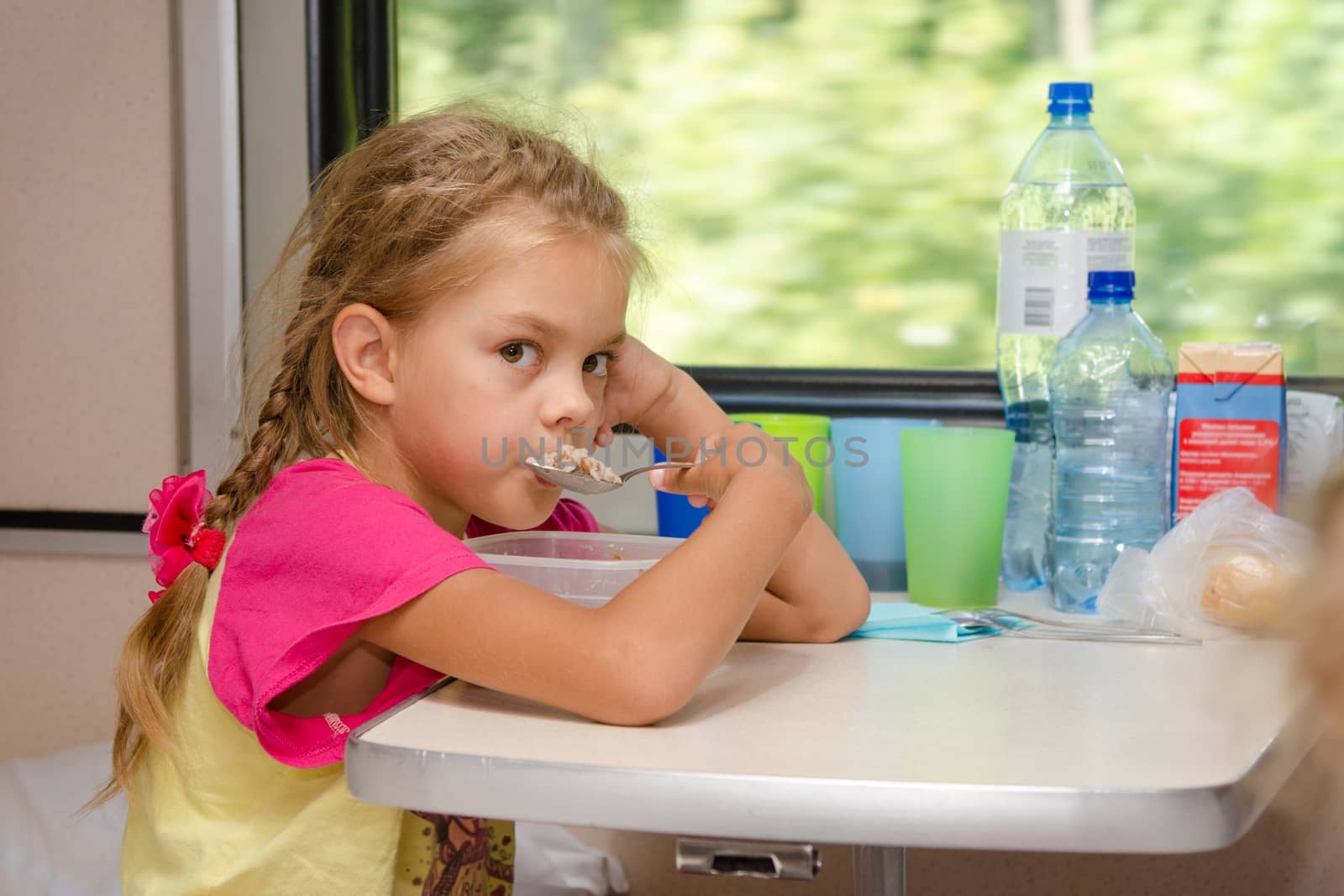 A six-year girl in a train sitting at the table on the lower place in the second-class compartment of the car and eats porridge spoon