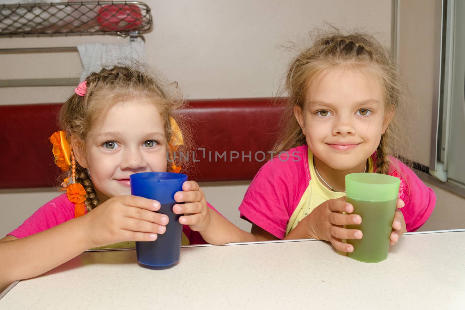 Two girls sisters on the train sitting at the table on the lower place in the second-class compartment wagon with glasses in hand by Madhourse