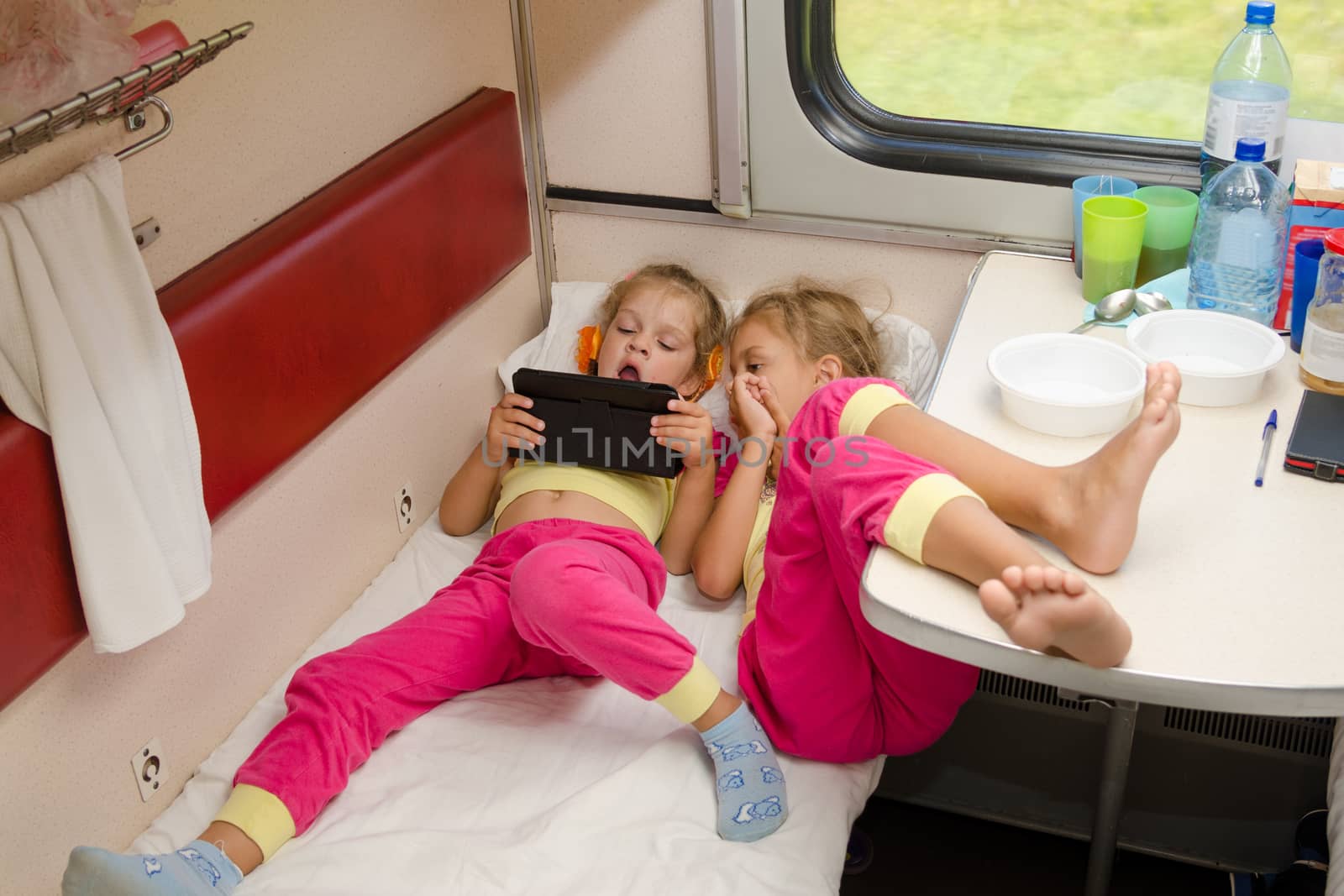 Two little girls lie on the train with his feet on the lower second-class place car in the same pajamas and looking at tablet