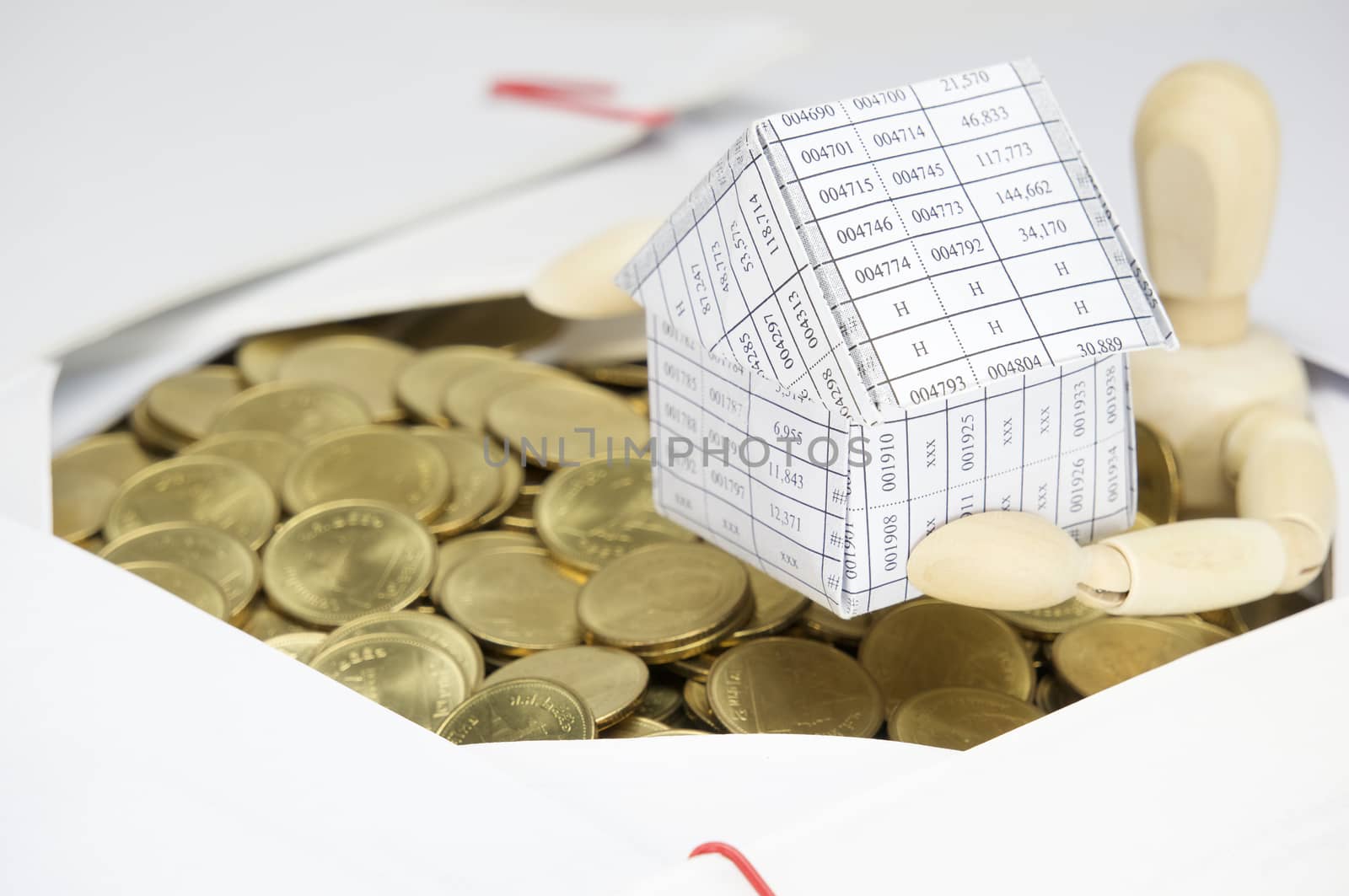 Close up wooden dummy holding house have surrounded by stack of gold coins and overload of paperwork as background.