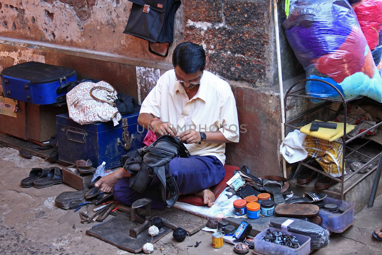 A man repairs rucksack on the footpath against building.Cobbler tools and necessities are around him. Vasco da Gama town, Goa, India.