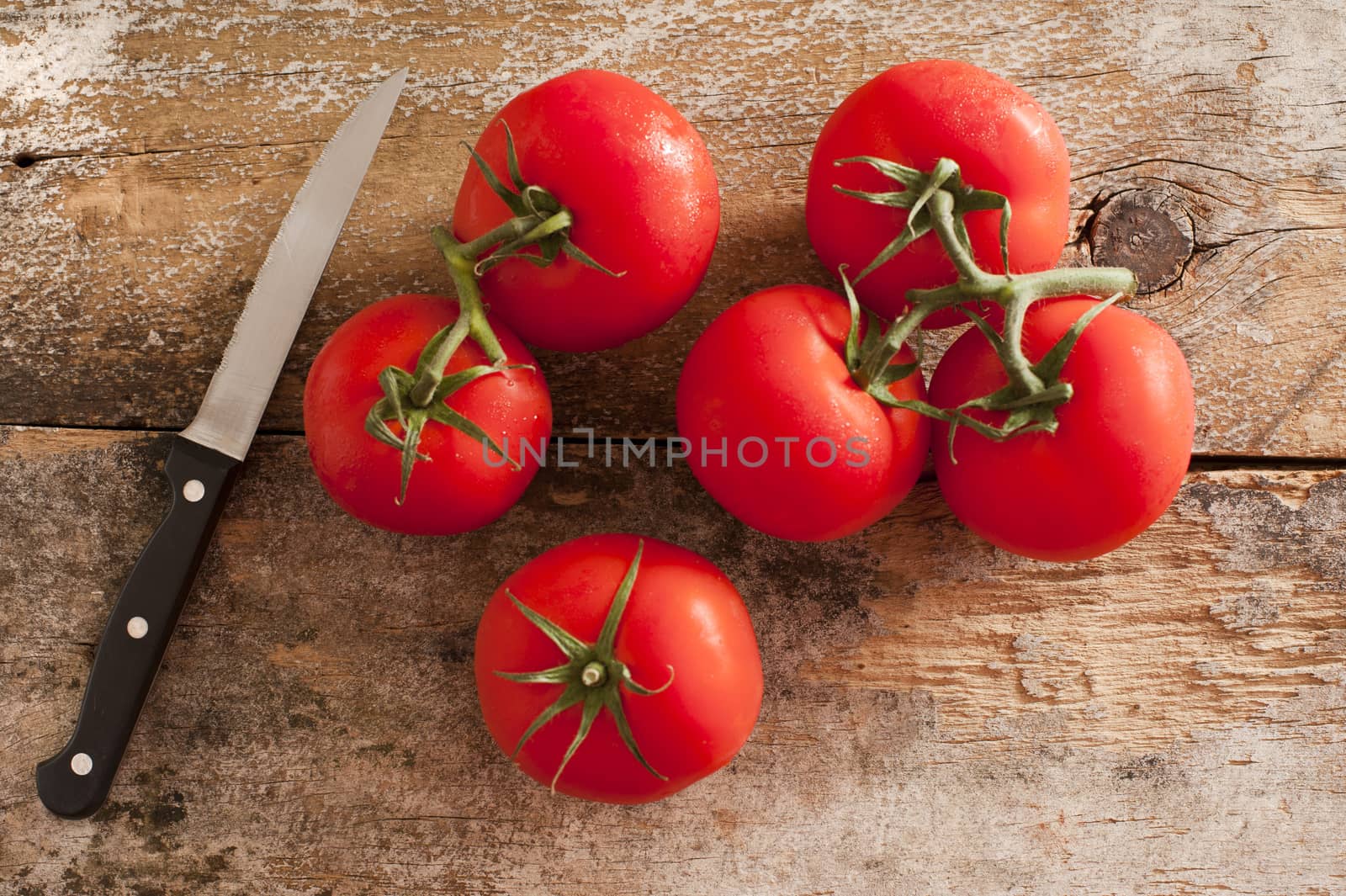 Six vine ripe red tomatoes with stems and single black and metal paring knife over weathered wooden table