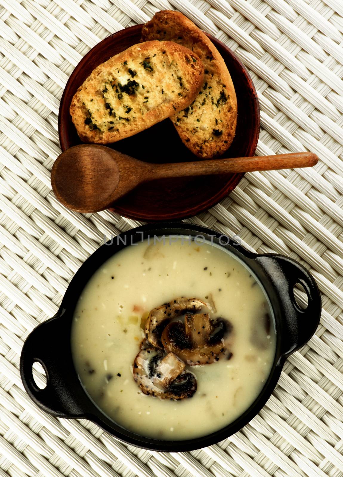 Delicious Homemade Mushrooms Cream Soup Decorated with Roasted Champignons in Black Iron Stewpot with Herb Crunchy Bread and Wooden Spoon closeup on Wicker background