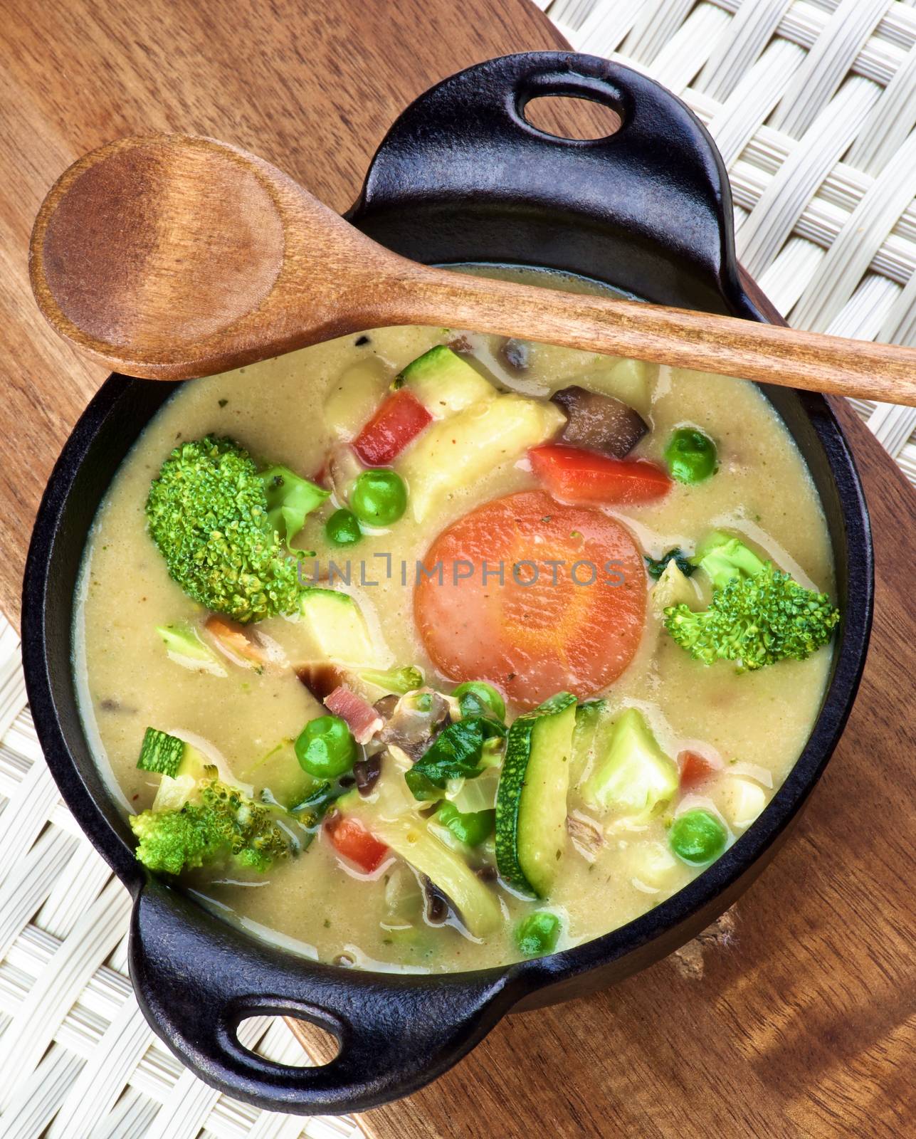 Rustic Vegetables Creamy Soup with Broccoli, Carrots, Zucchini, Leek, Red Bell Pepper and Green Pea in Black Iron Stewpot with Wooden Spoon closeup on Wooden Cutting Board. Top View