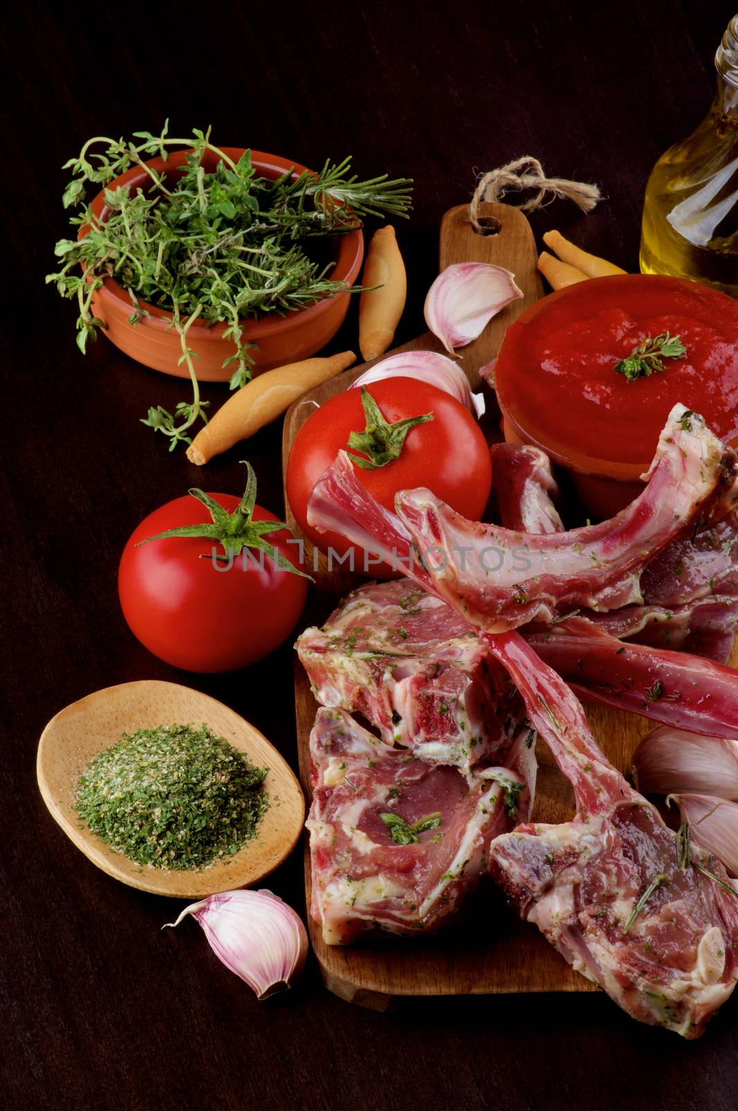 Arrangement of Raw Lamb Ribs with Tomatoes, Garlic, Herbs, Spices and Olive Oil on Wooden Cutting Board closeup on Dark Wooden background