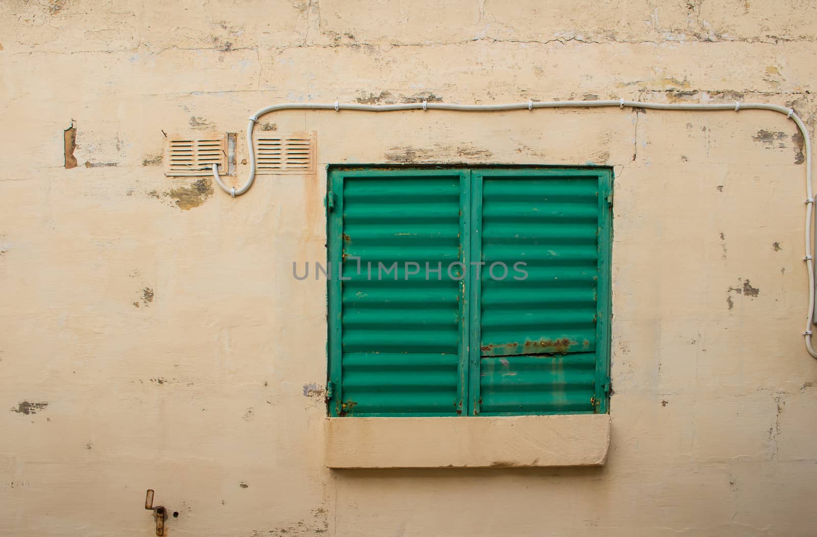 Old wall painted light yellow with a small window with a closed bright green shutter.