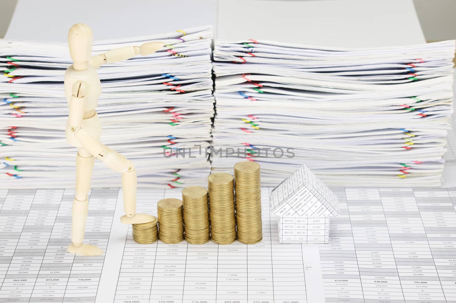 Wooden dummy step up with step pile of gold coins and house on finance account have blur overload of paperwork with colorful paperclip as background.