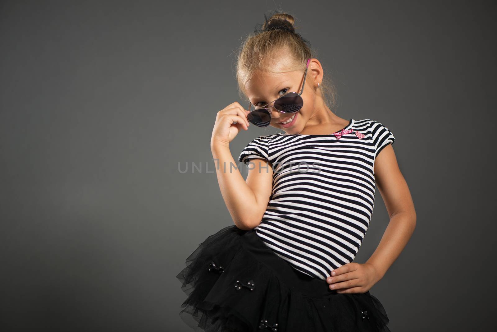 Pretty little girl smiling and posing with sunglasses. Studio shutting. Grey background. Looking at camera.