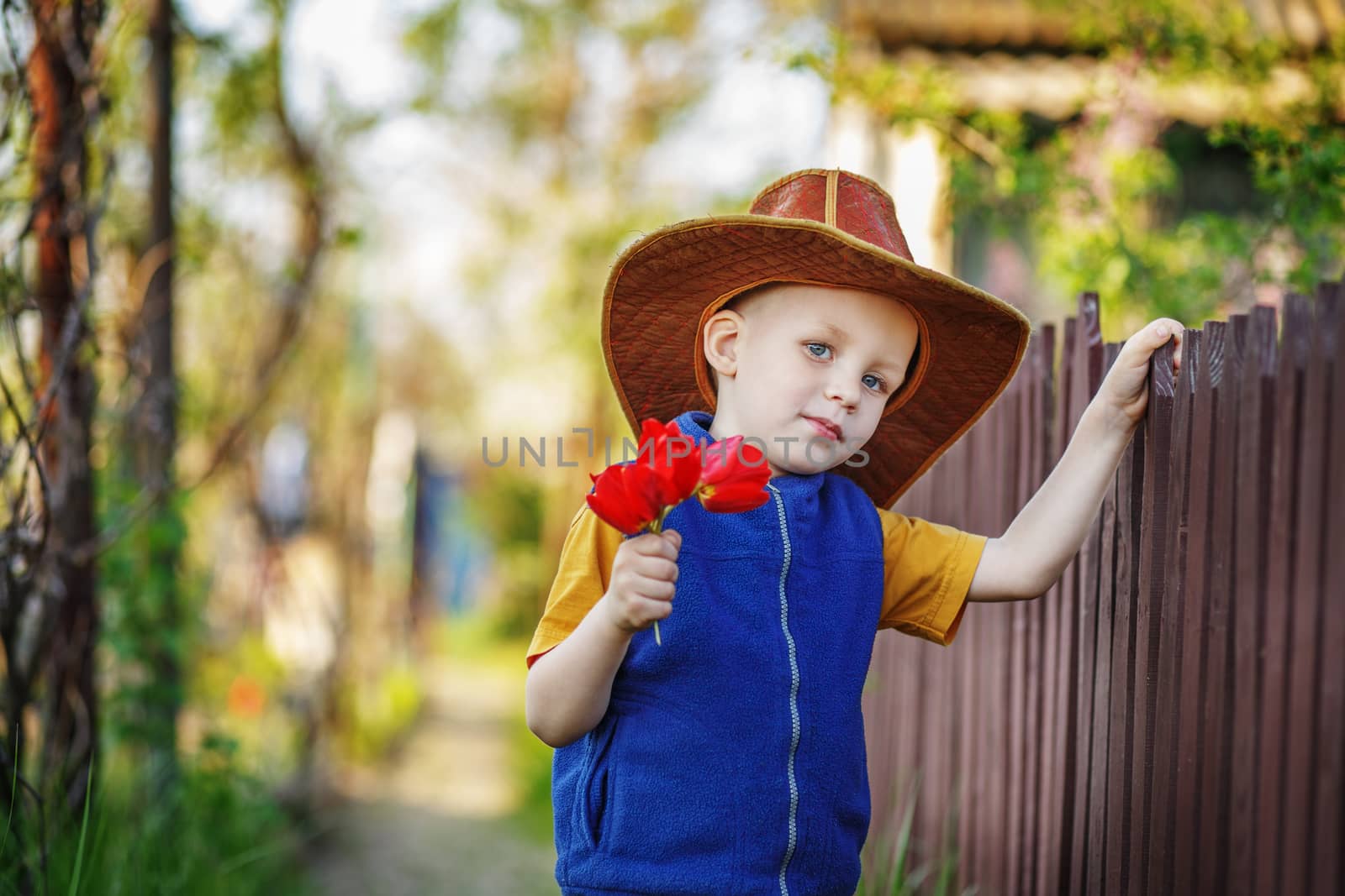 Portrait of a little boy standing in a big hat with a bouquet of tulips at the wooden fence in the countryside