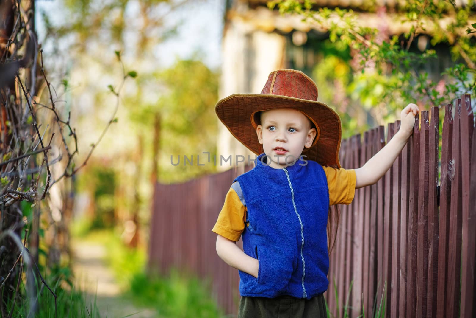 Portrait of a little boy standing in a big hat in the wooden fence in the countryside