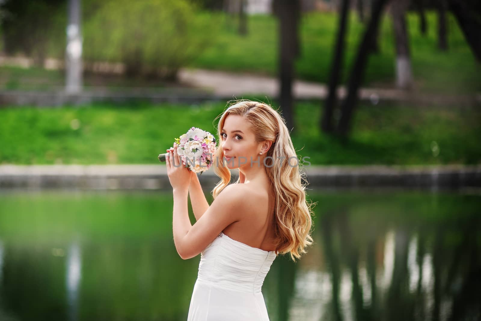 Young beautiful bride in a white dress with a bouquet standing near a pond waiting for the groom