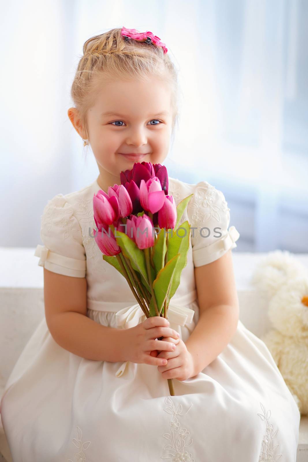 beautiful little girl in a white dress sitting on a windowsill with a bouquet of pink tulips