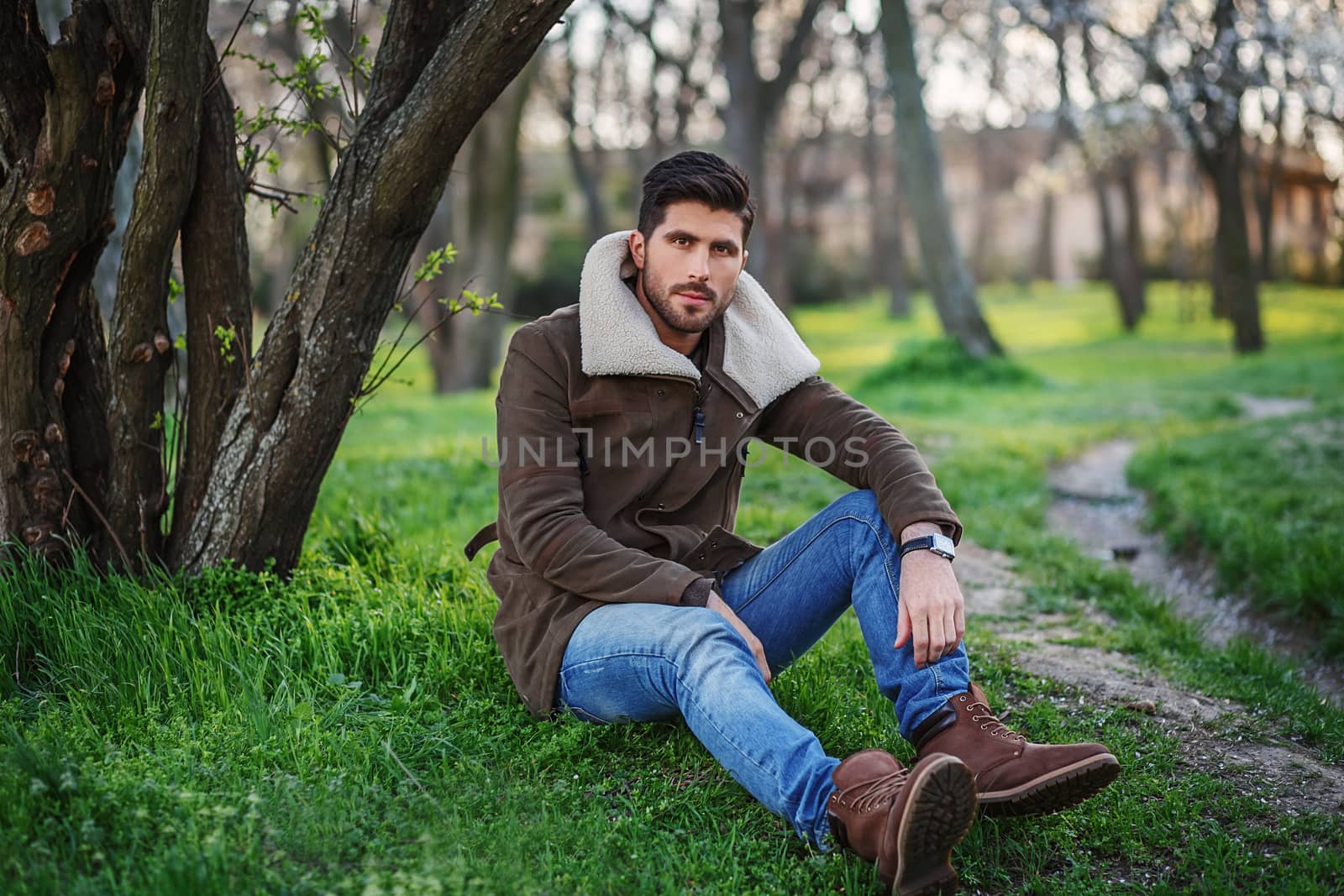 Portrait of trendy young attractive man sitting on green grass in a grove at sunset