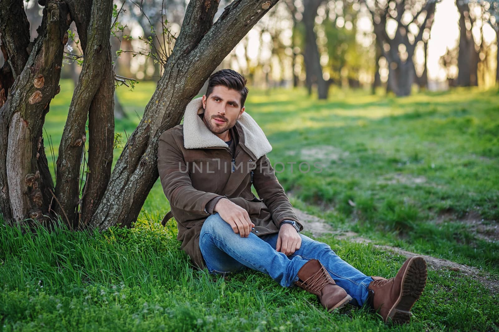 Portrait of trendy young attractive man sitting on green grass in a park at sunset