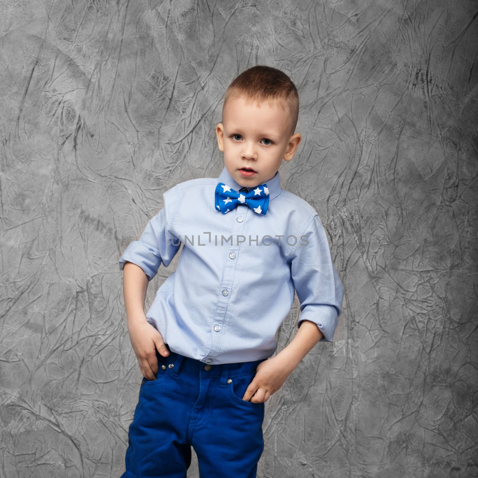 Portrait of a cute little boy in jeans, blue shirt and bow tie on a gray textural background in studio