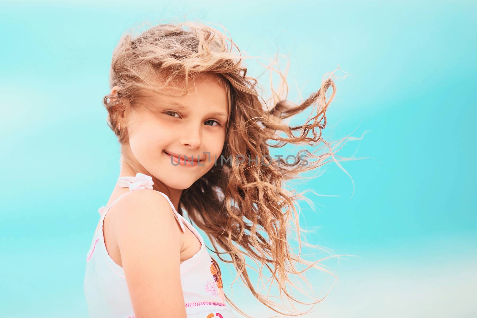 Portrait of a pretty little girl with waving in the wind long hair sitting on the beach against the blue sky