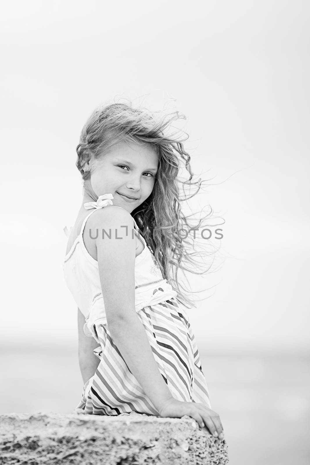 Portrait of a pretty little girl with waving in the wind long hair sitting on the beach. Black and white photo